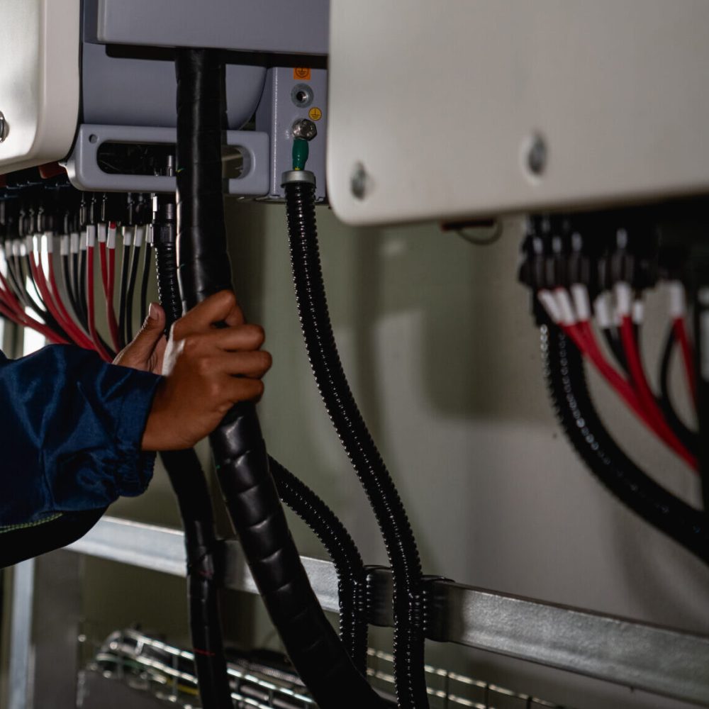 A woman in a yellow and blue safety vest is working on a power line. She is wearing a hard hat and is focused on her task